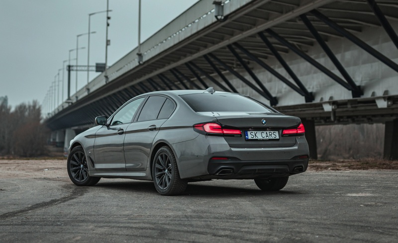 Задний вид BMW 5 серии серого цвета на фоне урбанистического пейзажа. Элегантный седан для аренды в SK CARS, идеально подходящий для любых поездок.