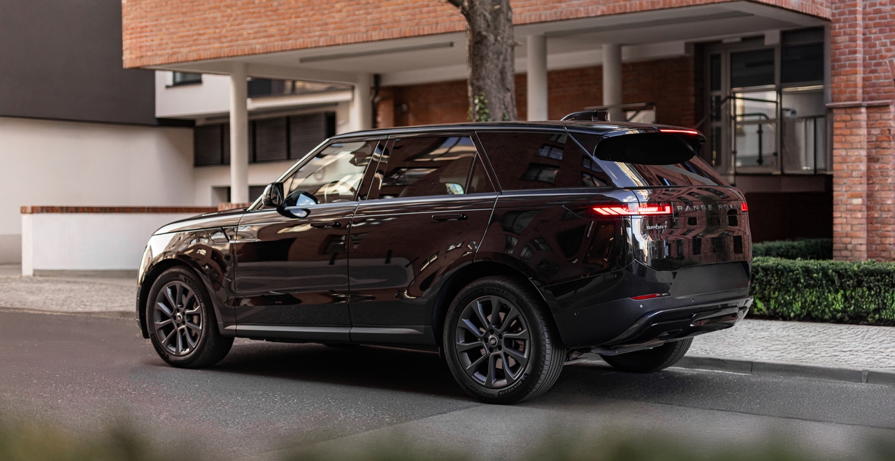 Чёрный Range Rover Sport, вид сзади под углом, припаркован на фоне кирпичного дома и современного фасада. Отражения зданий чётко видны на кузове автомобиля.