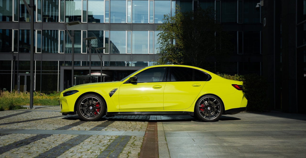 Ярко-жёлтый BMW M3 Competition xDrive на фоне современного офисного здания, вид сбоку.