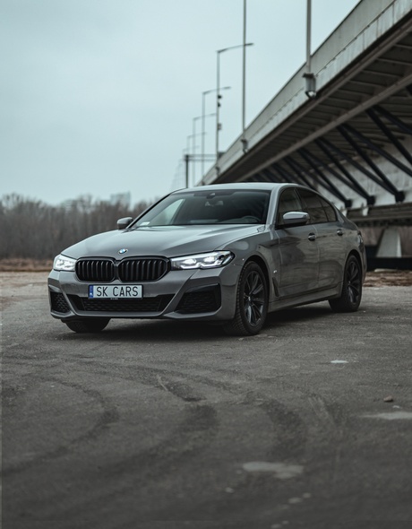 BMW 5 серии серого цвета на фоне урбанистического пейзажа под мостом. Стиль и мощь, доступные для аренды в SK CARS, идеальный выбор для городских поездок.