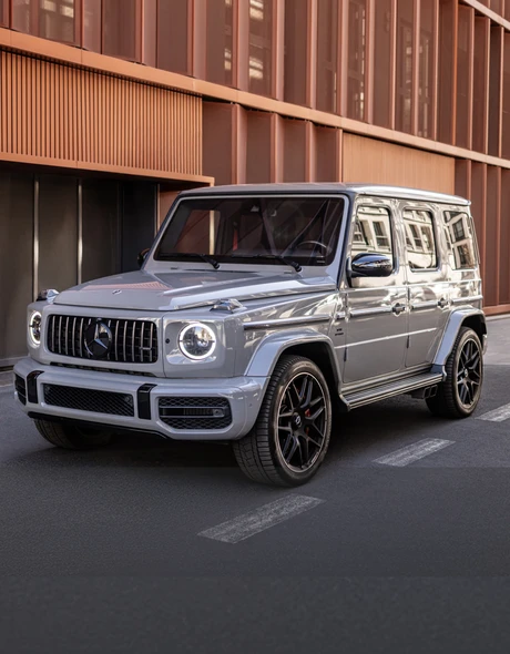 Mercedes G63 AMG на фоне современной архитектуры в центре города, вид спереди с включёнными дневными огнями.