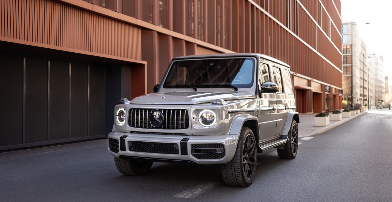 Mercedes G63 AMG на фоне современной архитектуры в центре города, вид спереди с включёнными дневными огнями.